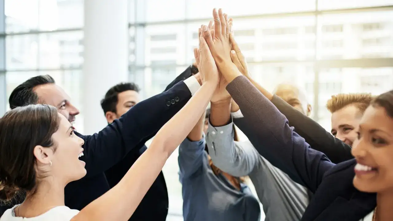 Un grupo diverso de profesionales celebra un &eacute;xito, uniendo sus manos en un gesto de unidad. Son la viva imagen de high performance teams.